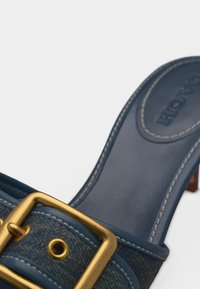 Κοντινό πλάνο μπλε δερμάτινου και τζιν σανδάλι mule με χρυσή πόρπη και εμφανές ράψιμο στην πάτημα και το λουρί.