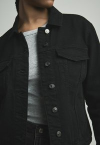 Zwarte denim jacket met zilverkleurige accenten, voorzien van een knoopsluiting aan de voorkant, twee borstzakken en een gestructureerde kraag over een grijs shirt.