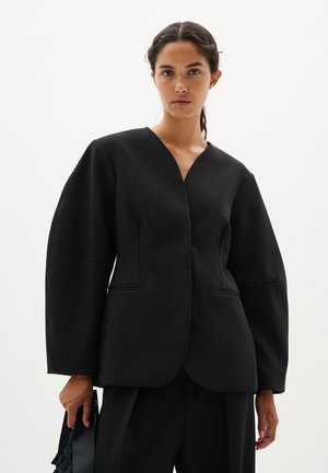Donna con capelli castani in un basso codino, veste un blazer nero strutturato e pantaloni abbinati, tiene una borsa nera contro uno sfondo semplice.