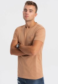 Lysebrun t-shirt med korte ærmer og rund halsudskæring samt en brystlomme. Stoffet ser glat ud med en subtil glans. Bæres med slidte denim.