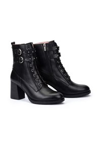 Botas de cuero negro hasta el tobillo con un tacón robusto, cordones frontales, cremallera lateral y correas ajustables con acentos metálicos. Forro interior suave.