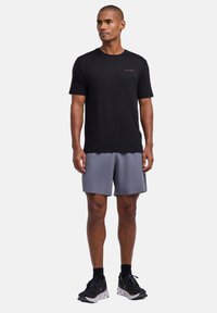 T-shirt noir à manches courtes avec un petit logo rouge, associé à un short gris. Le modèle porte des chaussures de sport, présentant un look décontracté de tenue de sport.