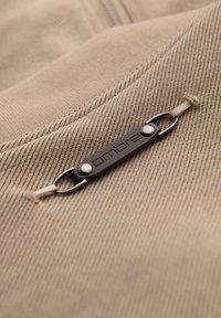 Beżowy materiał o żebrowej fakturze, z ciemnobrązową metką marki przymocowaną dwoma srebrnymi guzikami.