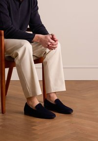 Marine blauwe suède instappers op blote voeten, gecombineerd met crèmekleurige broeken, zittend op een houten stoel op een visgraatvloer, met een ontspannen houding.