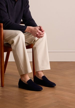Navy suede loafers on bare feet, paired with cream trousers, seated in a wooden chair on a herringbone floor, with relaxed posture.
