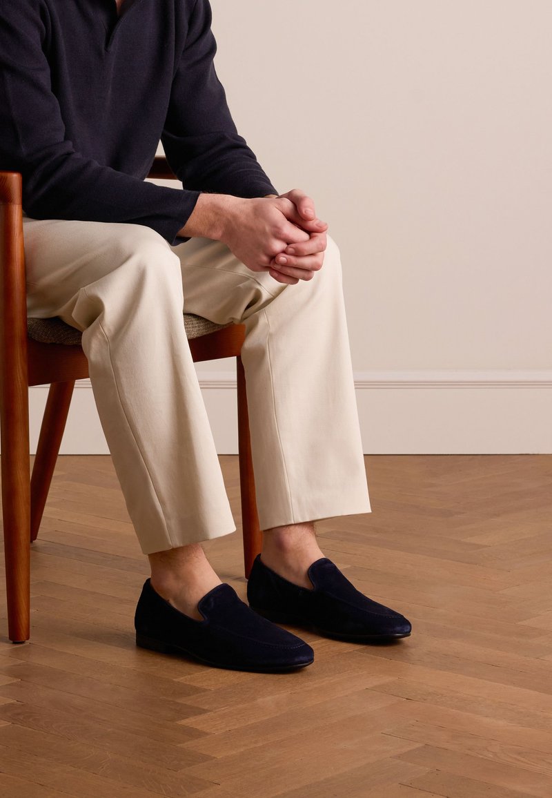 Persoon zit op een houten stoel met een gevlochten zitting, draagt marineblauwe loafers, beige broek en een marineblauw shirt met lange mouwen, handen gevouwen op schoot.