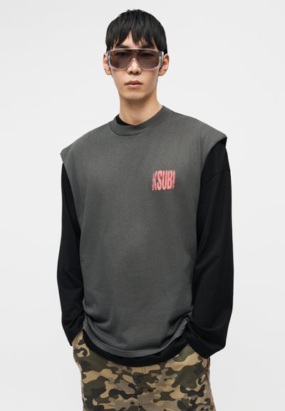Γκρι φαρδιά φούτερ με μαύρα μακριά μανίκια, διακοσμημένο με κόκκινο λογότυπο Ksubi. Συνδυασμένο με σορτς σε σχέδιο καμουφλάζ. Διαφανή γυαλιά ηλίου.