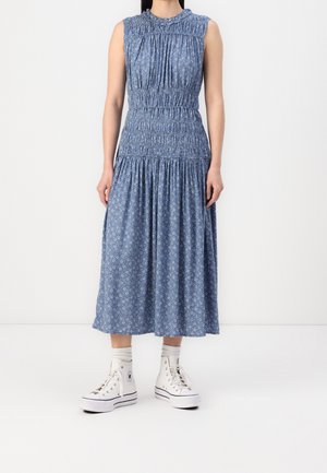 Mujer con vestido midi azul sin mangas con estampado, combinado con zapatillas blancas de caña alta y calcetines blancos, de pie sobre un fondo liso.