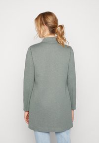 Manteau long vert menthe à la texture douce, col cranté et manches longues. Porté avec un jean bleu clair, présentant une coupe droite et un détail au dos.