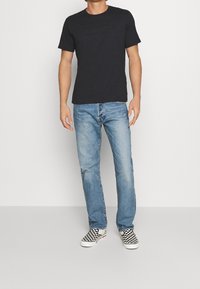 Osoba oblečená v černém tričku Levi's, světlých modrých džínách a černo-bílých kostkovaných slip-on botách, stojící na pozadí čistě bílé stěny.