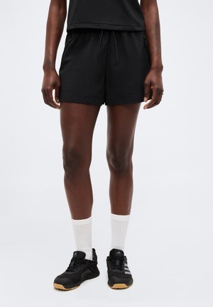 Personne portant un short de sport noir, un t-shirt noir, des chaussettes blanches et des chaussures de course noires, debout sur un fond clair.