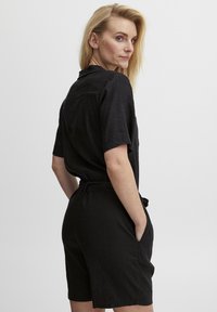 Juodas, trumpomis rankovėmis jumpsuitas su laisvu siluetu, turintis diržą talijoje ir kišenes. Pagamintas iš tekstūruoto, lengvo audinio.