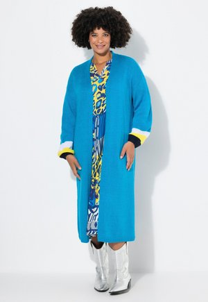 Un long cardigan tricoté bleu vif avec des poignets rayés blancs et jaunes, porté sur une robe à motif vif. Des bottes de cow-boy argentées complètent la tenue.