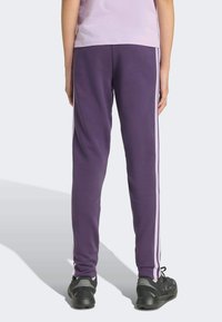 Fialové tepláky s bielymi bočnými pruhmi, vyrobené z mäkkého materiálu, s úzkym strihom. Styled s čiernymi športovými topánkami pre pridaný detail.