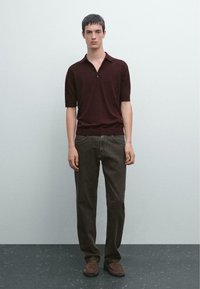Mörkbrun kortärmad polo med tre knappar, parat med mörkgrå rakbenade byxor och bruna mockaskor på en texturerad yta.
