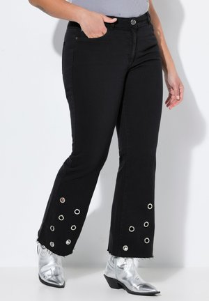 Zwarte flared jeans met een rafelrand en metalen oogjes bij de enkels. Gedragen met glanzende zilveren enkellaarsjes en een lichtgrijze top.