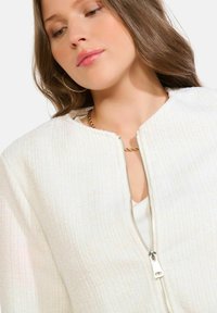 Balta zip-up jaka ar teksturētu audumu, kas piemirdz. Apaļš apkaklis, īsas piedurknes un redzama rāvējslēdzēja detaļa priekšpusē.