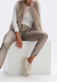 Femme portant un gilet en fourrure beige, une chemise blanche à manches longues, un pantalon taupe et des baskets blanches, assise sur un bloc blanc les jambes écartées.