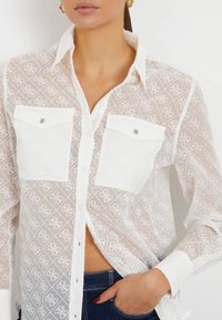 White, sheer button-up shirt with embroidered pattern, two front pockets with snap closures, and a collar. Suitable for layering.