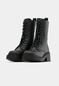 Μαύρες ψεύτικες δερμάτινες μποτές έως τον αστράγαλο με στρογγυλή μύτη, παχύ πέλμα και εμπρός κορδόνια. Διαθέτουν φερμουάρ στο πλάι και αντίθετη ράψιμο.