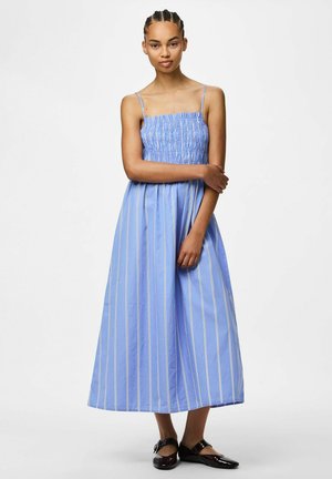 Jeune femme aux cheveux tressés portant une robe midi bleue sans manches à rayures verticales blanches et des chaussures Mary Jane noires, debout.