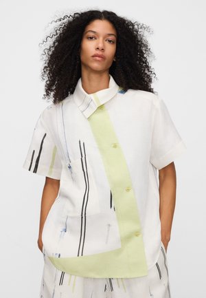 Femme aux cheveux bouclés portant une chemise à manches courtes et un pantalon assorti avec un motif à rayures abstraites blanches, vert pâle et noir.