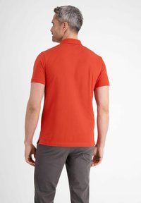 Czerwona bawełniana koszulka polo z krótkim rękawem i klasycznym kołnierzykiem, z bocznymi rozcięciami dla swobody ruchów. Noszona z szarymi spodniami.