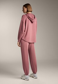 Přehnaně velká růžová mikina s kapucí, doplněná odpovídajícími zúženými tepláky. Materiál vypadá měkce s žebrovanými manžetami a bočními kapsami.