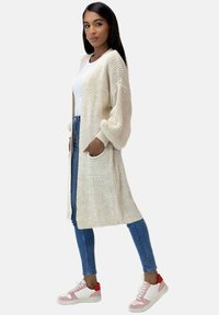 Lichtbeige gebreide cardigan met lange mouwen, open voorkant en twee voorzakken, gedragen over een witte top en blauwe jeans. Witte sneakers met roze accenten.