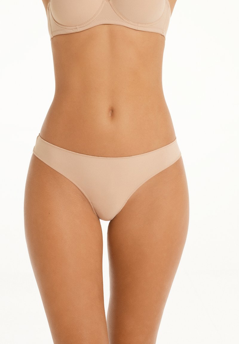Culotte en dentelle nude avec une texture lisse, un design taille basse et une couverture minimale sur les hanches, fabriquée dans un tissu doux et extensible.
