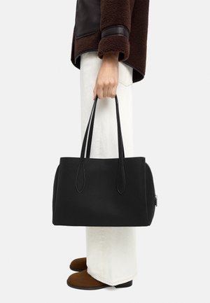 Osoba trzymająca dużą czarną torbę na ramię za uchwyty, ubrana w brązowe buty, białe spodnie oraz brązowo-czarną kurtkę, stojąca na białym tle.