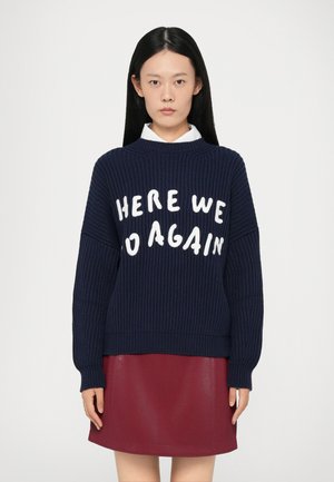 Maglione a maglia blu navy con maniche larghe, caratterizzato da audace scritta bianca "ECCOCI DI NUOVO." Abbinato a una gonna bordeaux lucida.