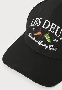 Căciulă neagră din țesătură, cu text alb brodat "LES DEUX", steaguri încrucișate și "International Sporting Goods" dedesubt.