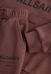 Nærbillede af brune sweatpants og sweatshirt med elastisk linning, ribbet cuff og sort "ALLSAINTS" tekst med japanske tegn.