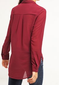 Burgundy long-sleeve shirt with a collar, side slits, and buttoned cuffs, made of a smooth, lightweight fabric. Features back pleat detail.