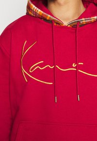 Sudadera roja con un logo bordado en dorado. Presenta un interior de capucha de cuadros, cordones ajustables y un bolsillo canguro en la parte delantera.