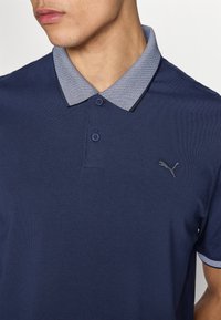 Μπλε σκούρο πόλο μπλουζάκι με υφή και σχεδιασμένο γιακά· διαθέτει δύο κουμπιά στον λαιμό και μικρό κεντημένο λογότυπο στο στήθος.