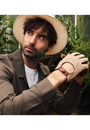 Man in beige wide-brimmed hat and brown jacket, wearing beige knit gloves with leather details, surrounded by green plants outdoors.