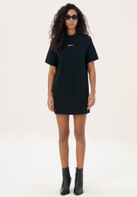 Zwarte oversized t-shirt jurk met korte mouwen, voorzien van een klein wit logo op de borst. Gecombineerd met zwarte enkellaarsjes.