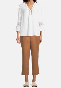 Blusa bianca a maniche lunghe con scollatura a V e dettagli ricamati ai polsi, abbinata a pantaloni marroni a gamba dritta e sandali chiari.