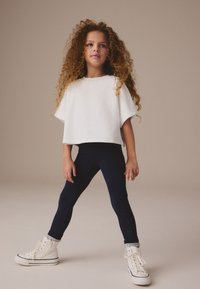 Valge üle mõõtmetega lühike topp, mereväe retuusid ja valged kõrge säärega tossud tekstuursete sokkidega; modell seisab neutraalsel taustal käed puusas.