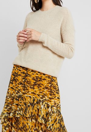 Femme en pull beige en tissu pelucheux et jupe à imprimé animal jaune, noir et blanc avec ourlet à volants, sur fond gris.