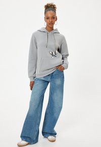 Sudadera gris con un diseño de perro bordado en el bolsillo, combinada con jeans de corte holgado en un tono azul claro y zapatos blancos.
