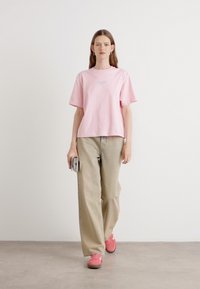 T-shirt in cotone rosa con maniche corte, vestibilità ampia; abbinata a pantaloni beige a gamba larga e scarpe rosa; in piedi su uno sfondo neutro.