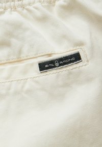 Beige tyg med en mjuk textur som har en svart "Sail Racing" logotagg sydd på sömmen. Lätta diagonala linjer synliga.