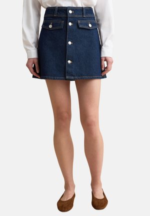 Mujer con minifalda vaquera azul oscuro con botones frontales y bolsillos con solapa, camisa blanca de manga larga y zapatos planos marrones.