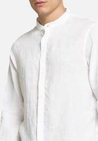 Camicia di lino bianca con colletto alla mandarino, chiusura frontale con bottoni e tessuto leggermente texturizzato. Vestibilità sartoriale con maniche lunghe.