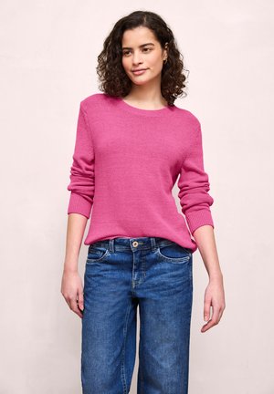 Femme aux cheveux bouclés et foncés portant un pull rose à manches longues et un jean bleu, debout devant un fond neutre clair.
