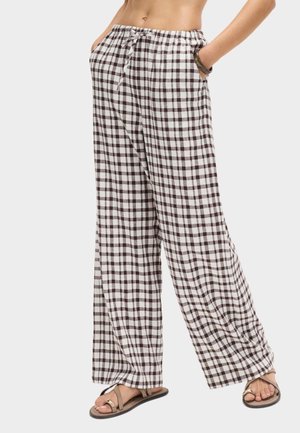 Femme portant un pantalon ample taille haute à carreaux noirs et blancs avec poches, associé à des sandales beiges à bout ouvert et un bracelet.