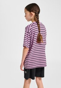 Lila gestreiftes kurzärmeliges T-Shirt aus weichem Stoff. Kombiniert mit schwarzen Shorts, die am Saum ein dekoratives Logo aufweisen.